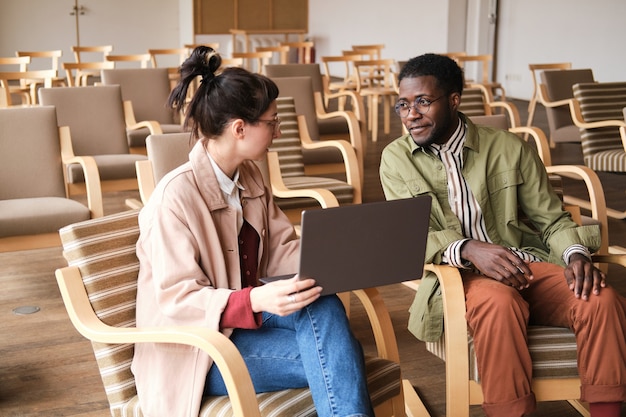 Young businesswoman using laptop and talking to her colleague while they sitting in empty conference hall