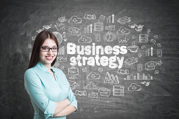 Young businesswoman wearing glasses and a blueish shirt standing with crossed arms and smiling. blackboard with a business strategy sketch