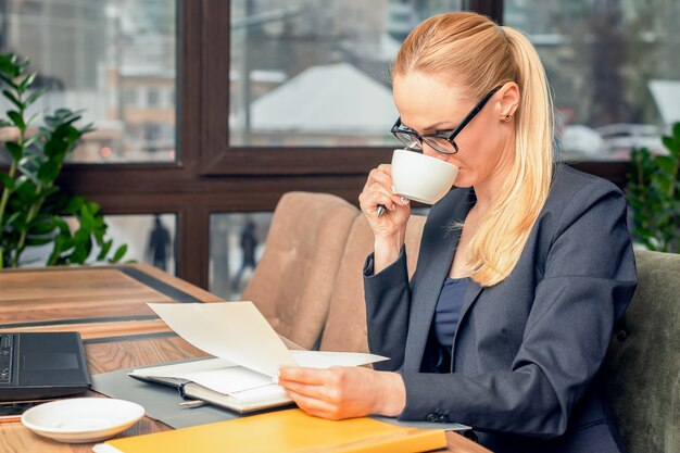 Young businesswoman wearing glasses sitting in coffee shop at table reads documents in front of laptop and drinking coffee.