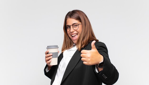 Молодая деловая женщина с кофе, чувствуя себя гордой, беззаботной, уверенной и счастливой, позитивно улыбаясь, подняв палец вверх