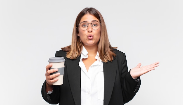 Giovane donna di affari con un caffè che sembra sorpreso e scioccato, con la mascella caduta tenendo un oggetto con una mano aperta sul lato