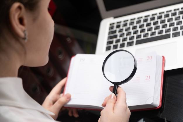 Giovane donna di affari con la lente d'ingrandimento che esamina diario sul fondo del computer portatile