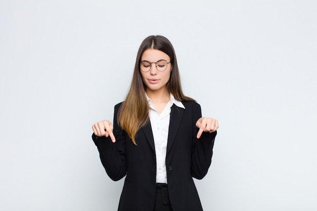 Young businesswoman with open mouth pointing downwards with both hands, looking shocked, amazed and surprised against white wall