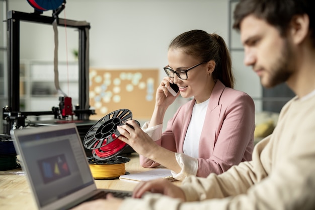 Young businesswoman with smartphone consulting one of clients about color and type of filament while sitting next to her co-worker