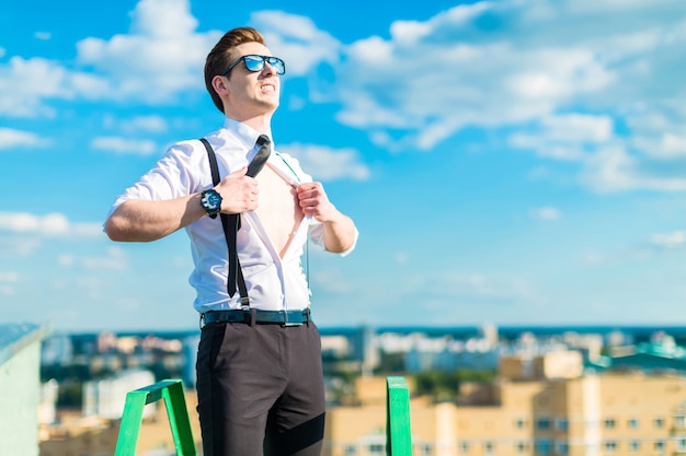 Young busunessman in white shirt, tie, braces and sunglasses stand on the roof ladder