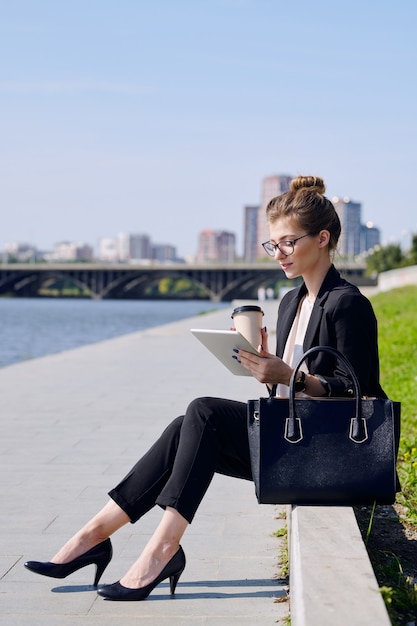 Young busy blond female in formalwear having drink and using tablet while sitting in front of riverside in urban environment on sunny day