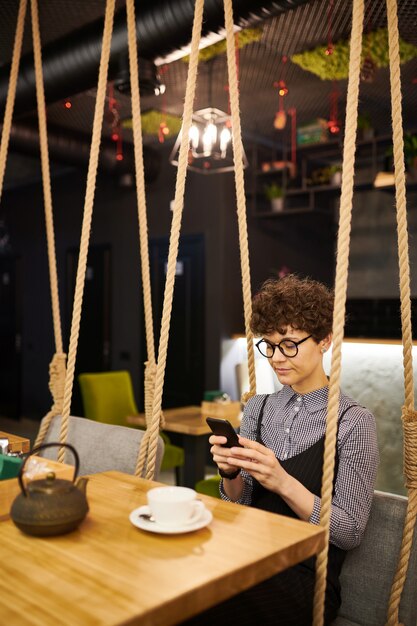 Young busy brunette woman scrolling in smartphone while sitting by table in comfy cafe by cup of tea