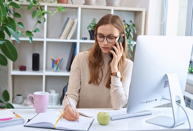 Young busy businesswoman attractive female employee in glasses sitting at desk talking by phone and