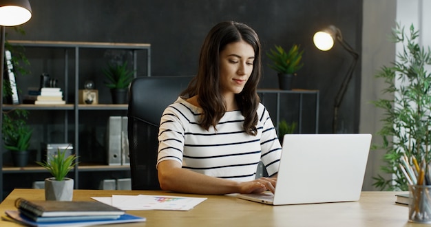 Young busy female office worker sitting at table, working on laptop computer and considering.