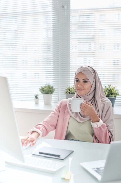 Young busy Muslim female designer or manager in hijab having tea or coffee while sitting by desk between window and computer monitor