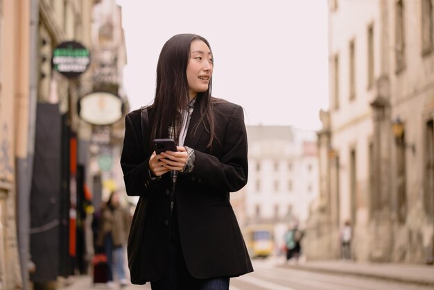 Photo young busy successful beautiful asian business woman korean professional businesswoman holding cellphone using smartphone standing or walking on old city street outside