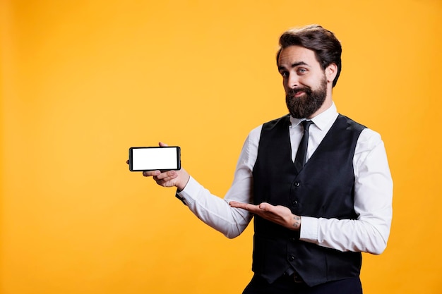 Young butler shows white display on camera, holding smartphone with isolated copyspace screen. Professional skilled restaurant employee presents blank empty screen template in studio.