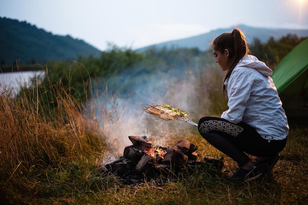 Young calm woman tourist frying vegetables at the beautiful nature landscape while sitting near the tent by the fire Travel tourism camping concept