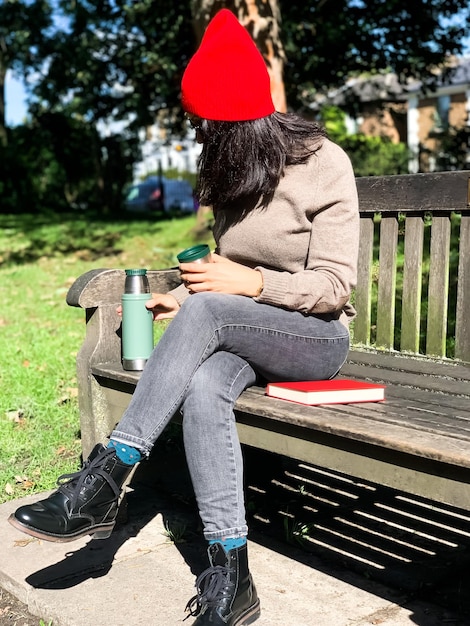 Foto giovane donna millenaria in abiti casuali d'autunno seduta nel parco e che beve caffè da un thermos