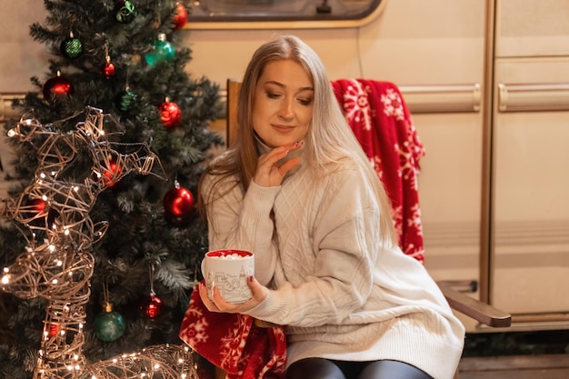 Giovane donna candida in maglione accogliente lavorato a maglia che tiene in mano una tazza di bevanda al cacao con marshmallow seduto rilassante sulla sedia campeggio all'aperto e godimento felice anno nuovo e buon natale vigilia