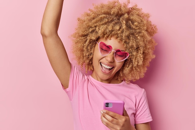 Young carefree curly woman makes triumph dance keeps arm raised\
up laughs gladfully uses mobile phone for chatting online wears\
heart shaped sunglasses and t shirt isolated over pink\
background