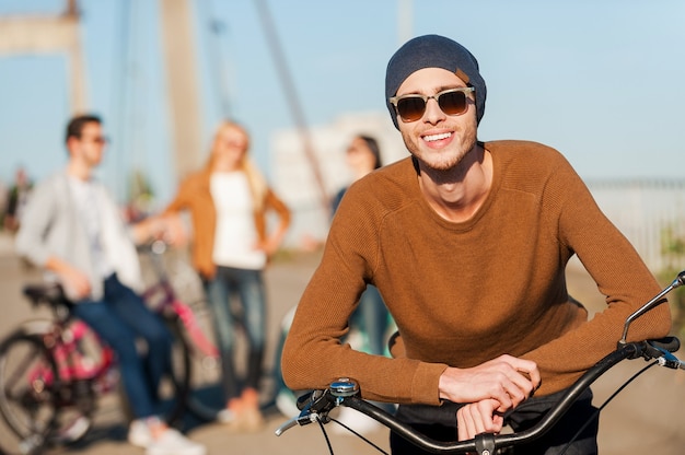 若くてのんき。彼の友人がバックグラウンドで話している間、彼の自転車に寄りかかって、笑顔でカメラを見ているハンサムな若い男