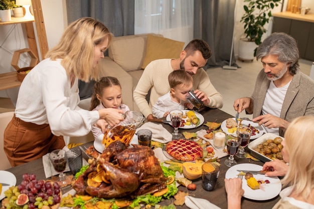 Photo young careful parents giving drinks to their cute little son and daughter by served festive table during thanksgiving dinner with grandparents