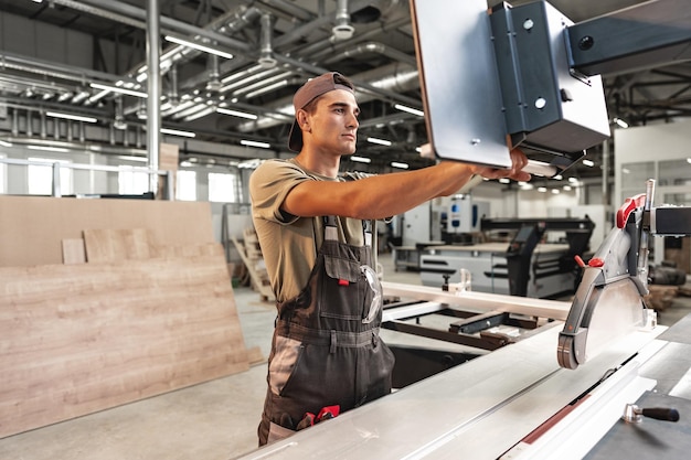 Young carpenter cutting a piece of wood in using a circular saw in furniture factory close up