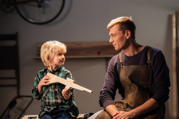 Foto un giovane papà falegname e il suo grazioso figlio biondo hanno costruito una pistola di legno e lo dimostrano, sono felici, seduti nella bottega del falegname sul tavolo.