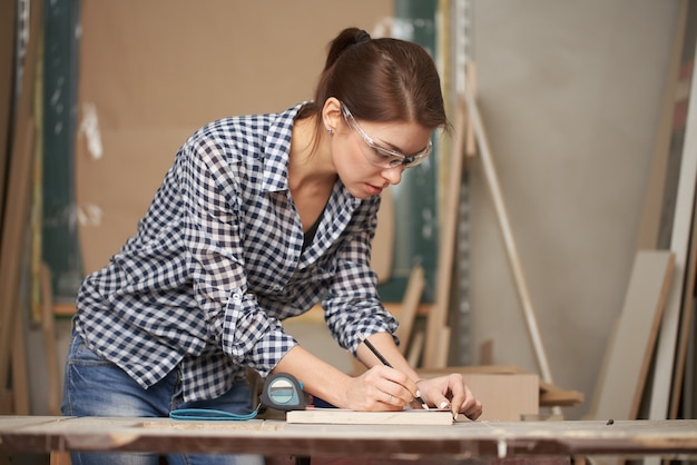 Young carpenter in glasses with tape measure and blackboard in workshop