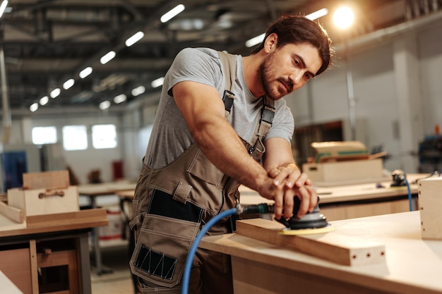 Young carpenter sanding wood piece in workshop in furniture factory close up