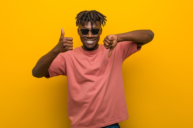 Young casual black man wearing sunglasses showing thumbs up and thumbs down, difficult choose concept