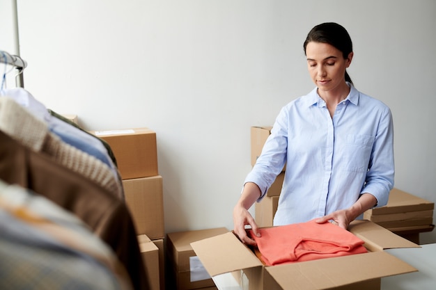 young casual employee putting folded casualwear into box in process of packing client order in office
