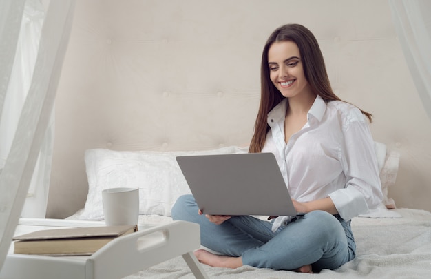 Young casual girl working on laptop from her bed during quarantine and smiling