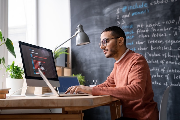 Young casual it-manager sitting in front of laptop in office and analyzing coded information