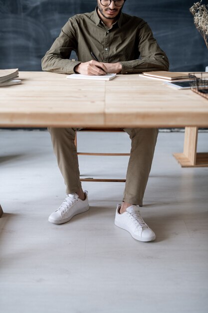 Photo young casual man sitting by wooden table in office or classroom and making notes in notebook