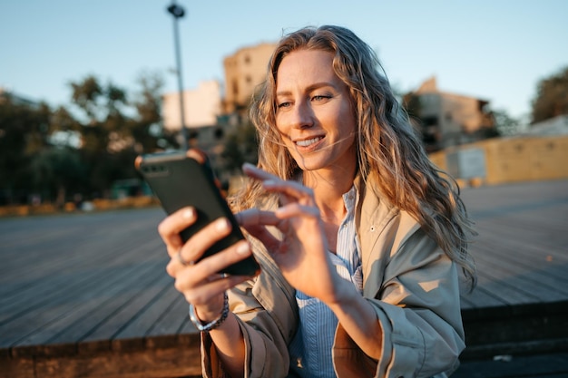 スマートフォンを使用して海の堤防のベンチに座っているコートを着た若いカジュアルな女性