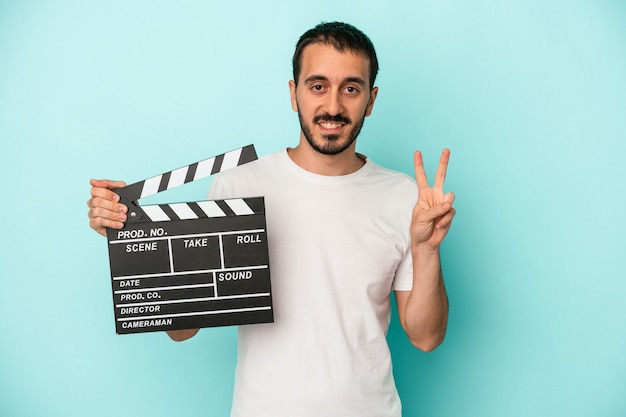 Young caucasian actor man holding clapperboard isolated on blue background showing number two with fingers.