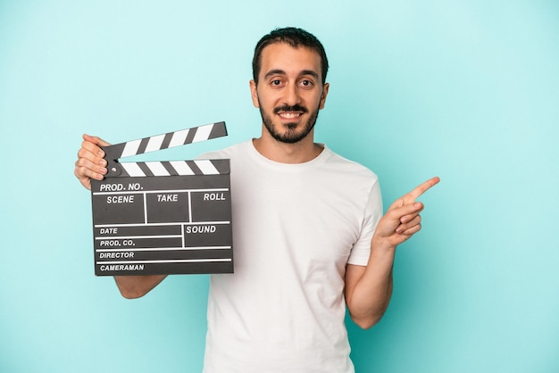 Young caucasian actor man holding clapperboard isolated on blue background smiling and pointing aside showing something at blank space