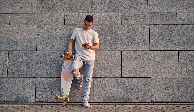 Young Caucasian American guy in glasses with a longboard against the gray wall of an urban building