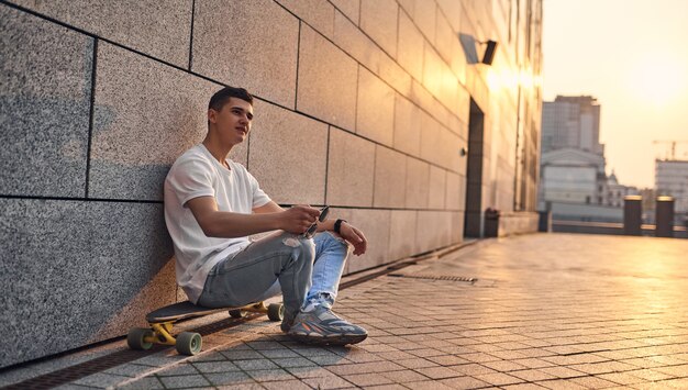 A young Caucasian American guy sits crosslegged on a longboard glasses in hand against the wall