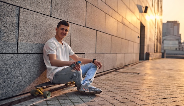 A young Caucasian American guy sits crosslegged on a longboard glasses in hand against the wall