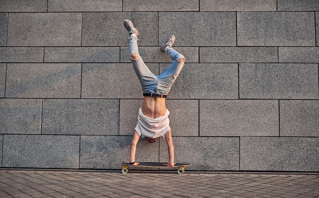 Photo a young caucasian american guy stands on his hands on a longboard against the gray wall of building