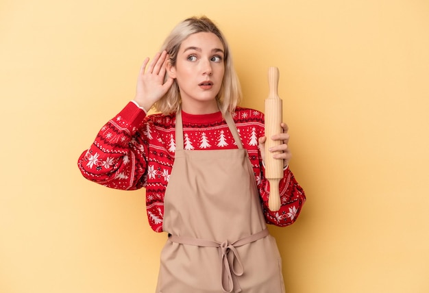 Young caucasian baker woman doing cookies for Christmas isolated on yellow background trying to listening a gossip.