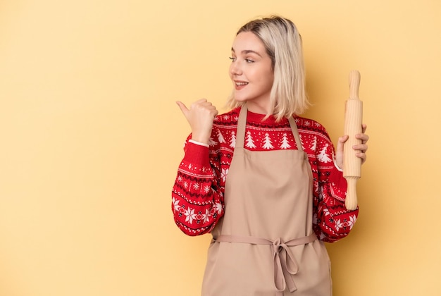 写真 離れて親指の指で黄色の背景のポイントに分離されたクリスマスのクッキーをやっている若い白人のパン屋の女性は、笑ってのんき。