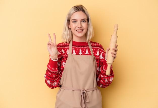 写真 指で2番目を示す黄色の背景に分離されたクリスマスのクッキーをやっている若い白人のパン屋の女性。