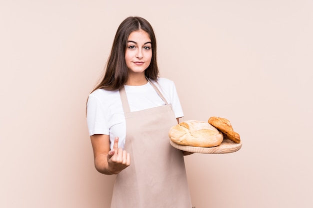 Young caucasian baker woman pointing with finger at you as if inviting come closer.