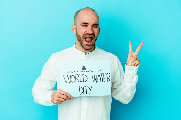 Young caucasian bald man celebrating world water day isolated on blue joyful and carefree showing a peace symbol with fingers.