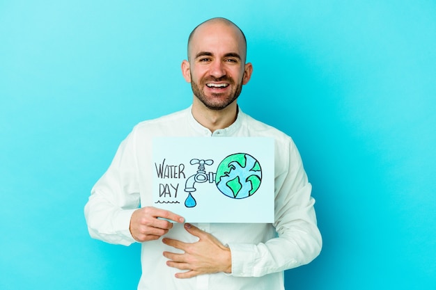 Foto giovane uomo calvo caucasico che celebra la giornata mondiale dell'acqua isolata sul blu ridendo e divertendosi.