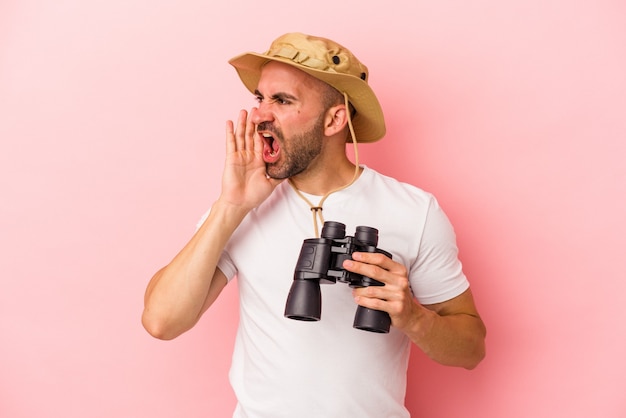 Young caucasian bald man holding binoculars isolated on pink background  shouting and holding palm near opened mouth.