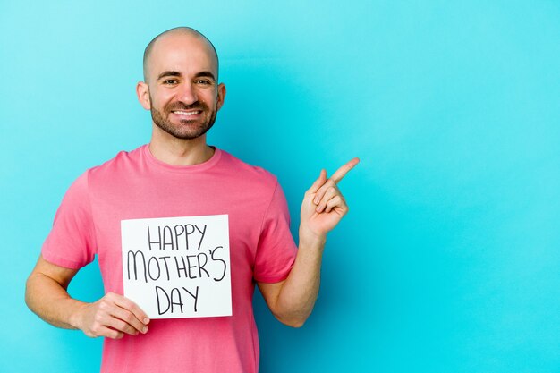 Молодой кавказский лысый мужчина держит плакат дня счастливой матери на синем, улыбаясь и указывая в сторону, показывая что-то на пустом месте.