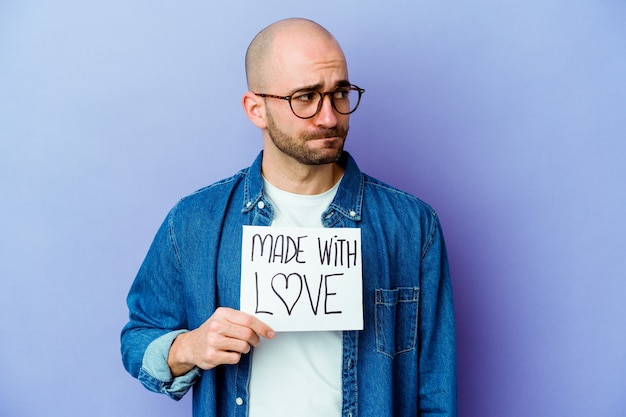 Young caucasian bald man holding a made with love placard isolated on purple wall confused, feels doubtful and unsure.