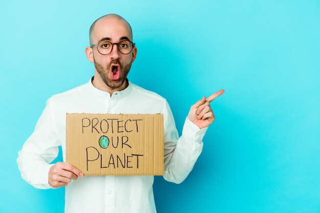 Young caucasian bald man holding a protect our planet placard isolated on purple background pointing to the side