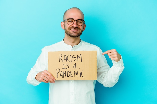 Молодой кавказский лысый мужчина, держащий в руках расизм, - пандемия, изолированный на белой стене, человек, гордый и уверенный, указывая рукой на место для копирования рубашки