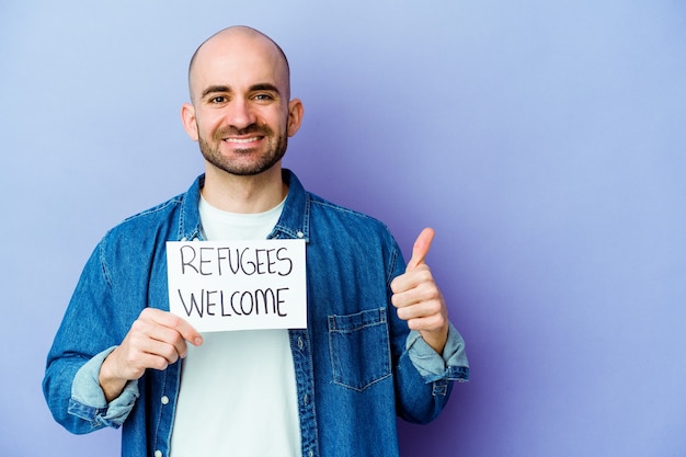 Young caucasian bald man holding a Refugees welcome placard isolated on blue background smiling and raising thumb up